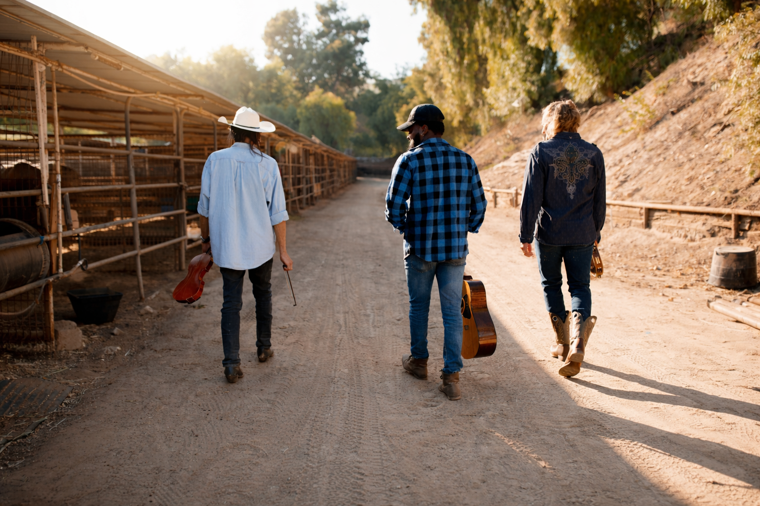 Tres músicos caminan por un camino de tierra en entorno rural de la zona central de Chile, con instrumentos y corrales al costado.