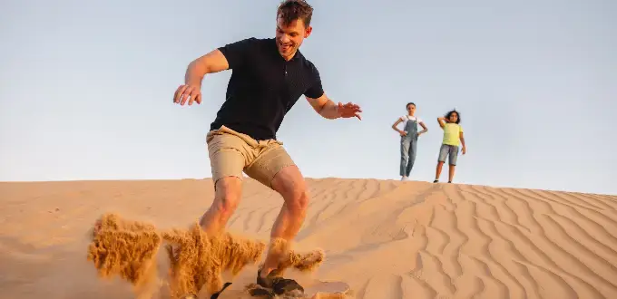 Hombre practicando sandboard en las Dunas de Reñaca, Viña del Mar, con personas caminando sobre la arena al atardecer en la costa central de Chile.