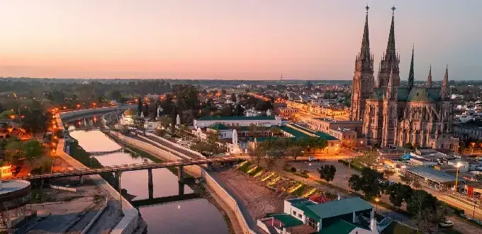 Panorámica de Luján, Buenos Aires, con la Basílica de Luján al atardecer y el río con puentes iluminados.
