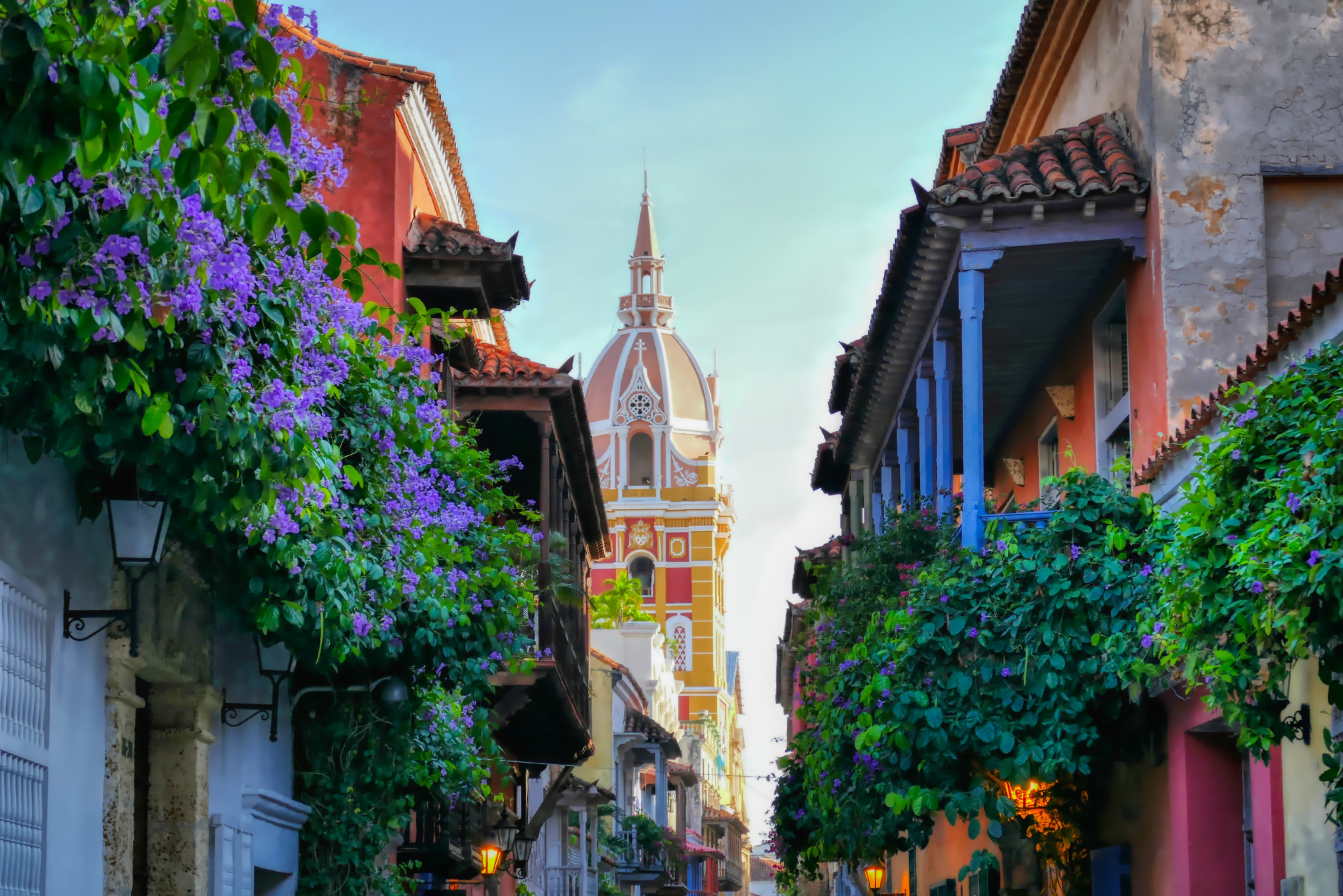 Calle colonial en el Centro Histórico de Cartagena, Colombia, con flores coloridas y la cúpula de la catedral al fondo.