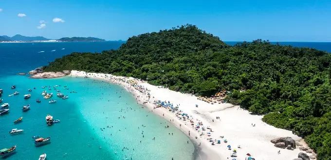 Playa de Florianópolis, Brasil, con aguas cristalinas, arena blanca, vegetación tropical y barcos en la costa.
