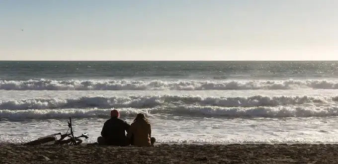 Pareja sentada frente al mar al atardecer, con olas del Pacífico y bicicleta en la arena, costa de Chile.