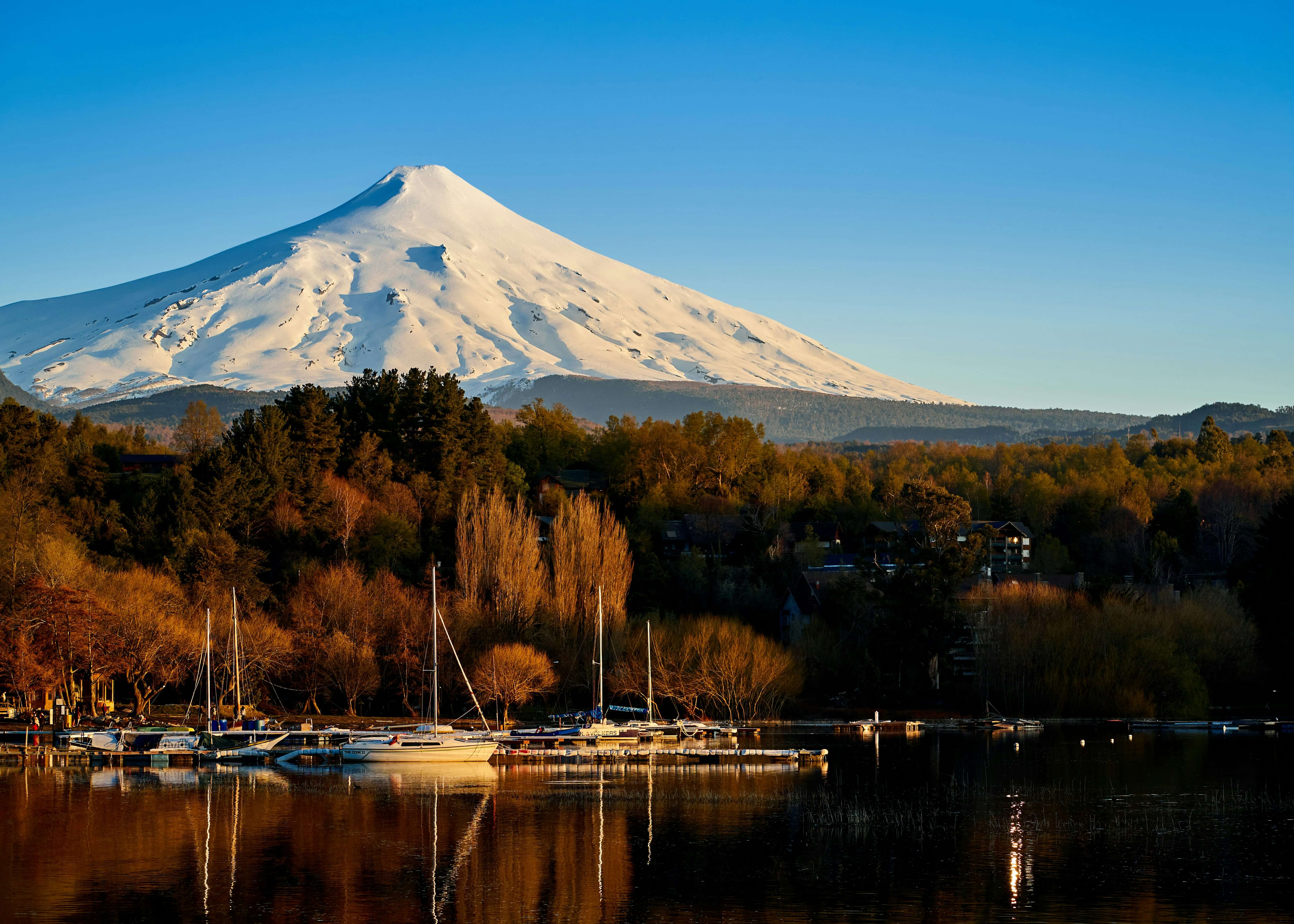 Volcán Villarrica nevado sobre el lago con veleros y bosque otoñal en Pucón, Región de La Araucanía, Chile.