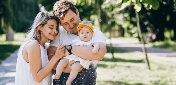 Familia joven con bebé disfrutando un paseo al aire libre en un parque urbano, rodeados de árboles y luz natural.