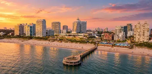 Vista aérea de Punta del Este, Uruguay, con playa, muelle, edificios costeros y un atardecer sobre el mar.