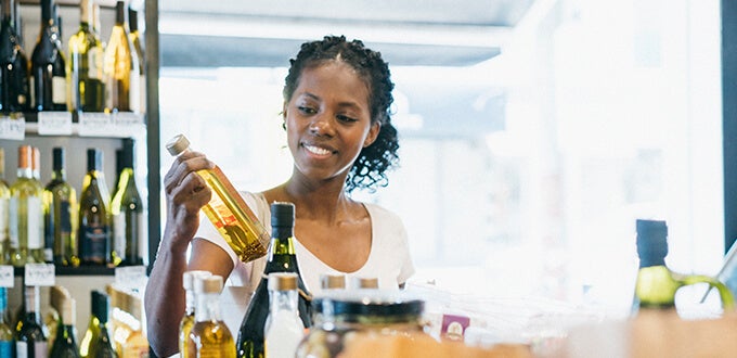 Mujer selecciona una botella de aceite en tienda de alimentos, rodeada de estantes con vinos y productos gourmet.