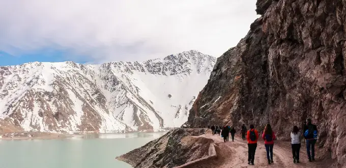 Sendero junto al Embalse El Yeso, con caminantes, montañas nevadas y aguas turquesas en el Cajón del Maipo, Chile.
