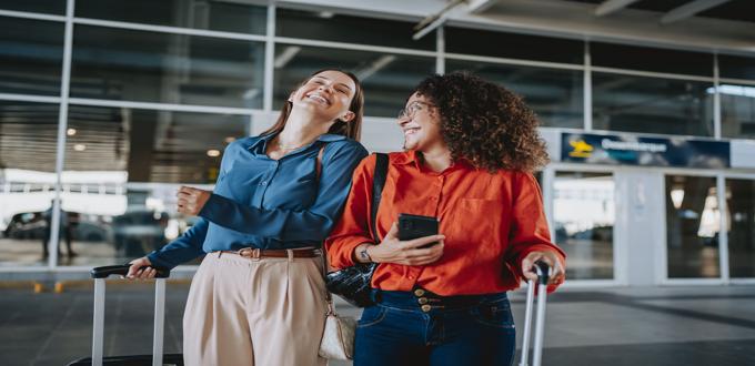 Dos mujeres riendo mientras caminan con sus maletas en el aeropuerto.