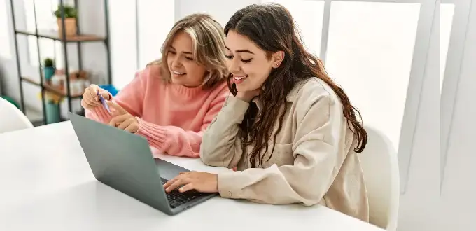 Dos amigas sonríen mientras usan un computador portátil y revisan notas en una mesa en Bellavista.