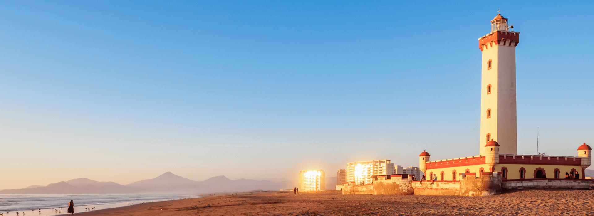Faro Monumental de La Serena al atardecer, junto a la playa, con edificios y montañas de fondo.