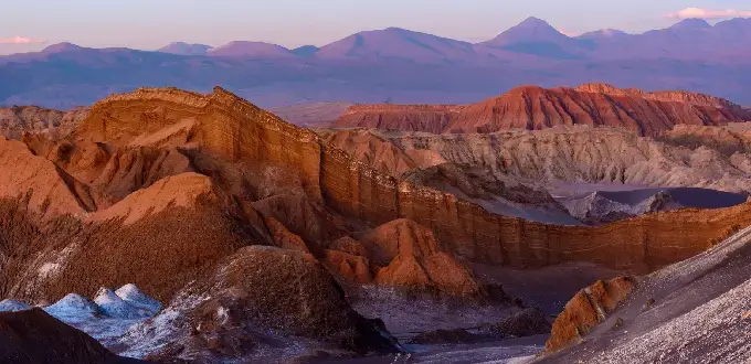 Formaciones rocosas y dunas del Valle de la Luna en San Pedro de Atacama, con cordillera y tonos rojizos al atardecer
