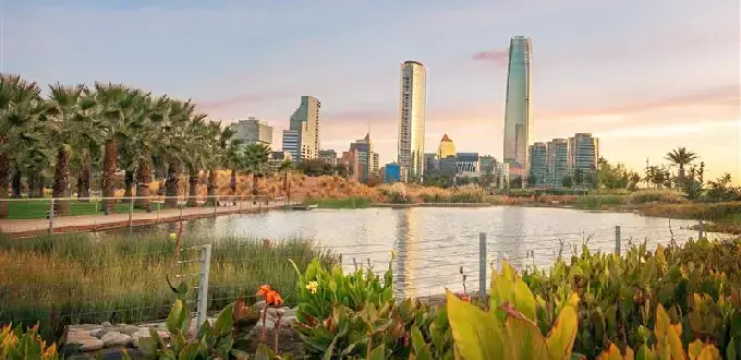 Laguna y áreas verdes del Parque Bicentenario en Vitacura con el skyline de Santiago y la Gran Torre Santiago al atardecer.