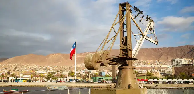Grúa histórica en puerto de Antofagasta con bandera de Chile, ciudad costera y cerros del desierto de Atacama al fondo