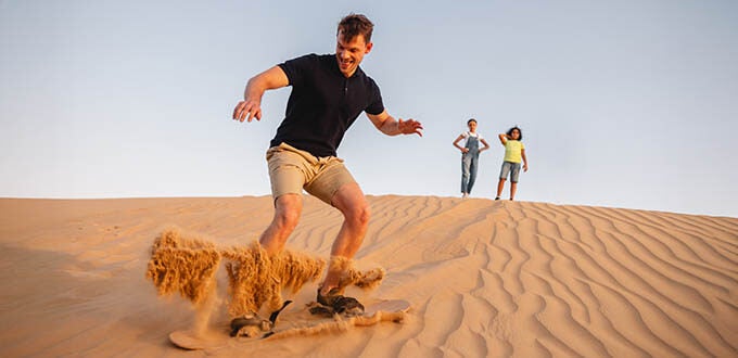 Hombre practicando sandboard en dunas del desierto con familia al fondo, aventura turística en entorno árido.
