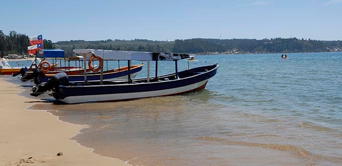 Embarcaciones turísticas varadas en la orilla, playa tranquila y aguas calmas con colinas al fondo, sur de Chile.