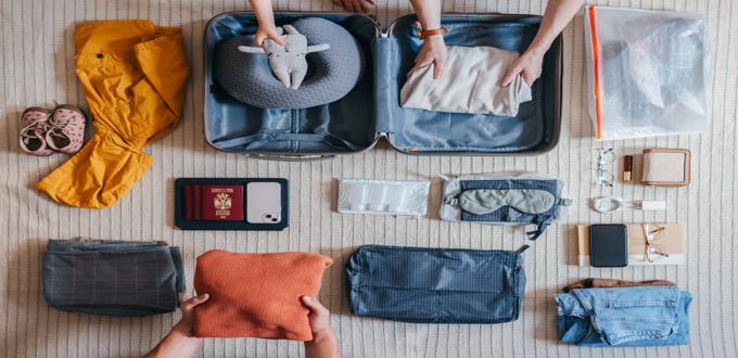 Persona empacando una maleta con ropa, accesorios de viaje y documentos organizados sobre una alfombra, vista desde arriba.