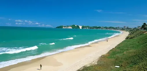 Playa de arena blanca y aguas turquesas en Natal, Brasil, rodeada de dunas y vegetación costera.
