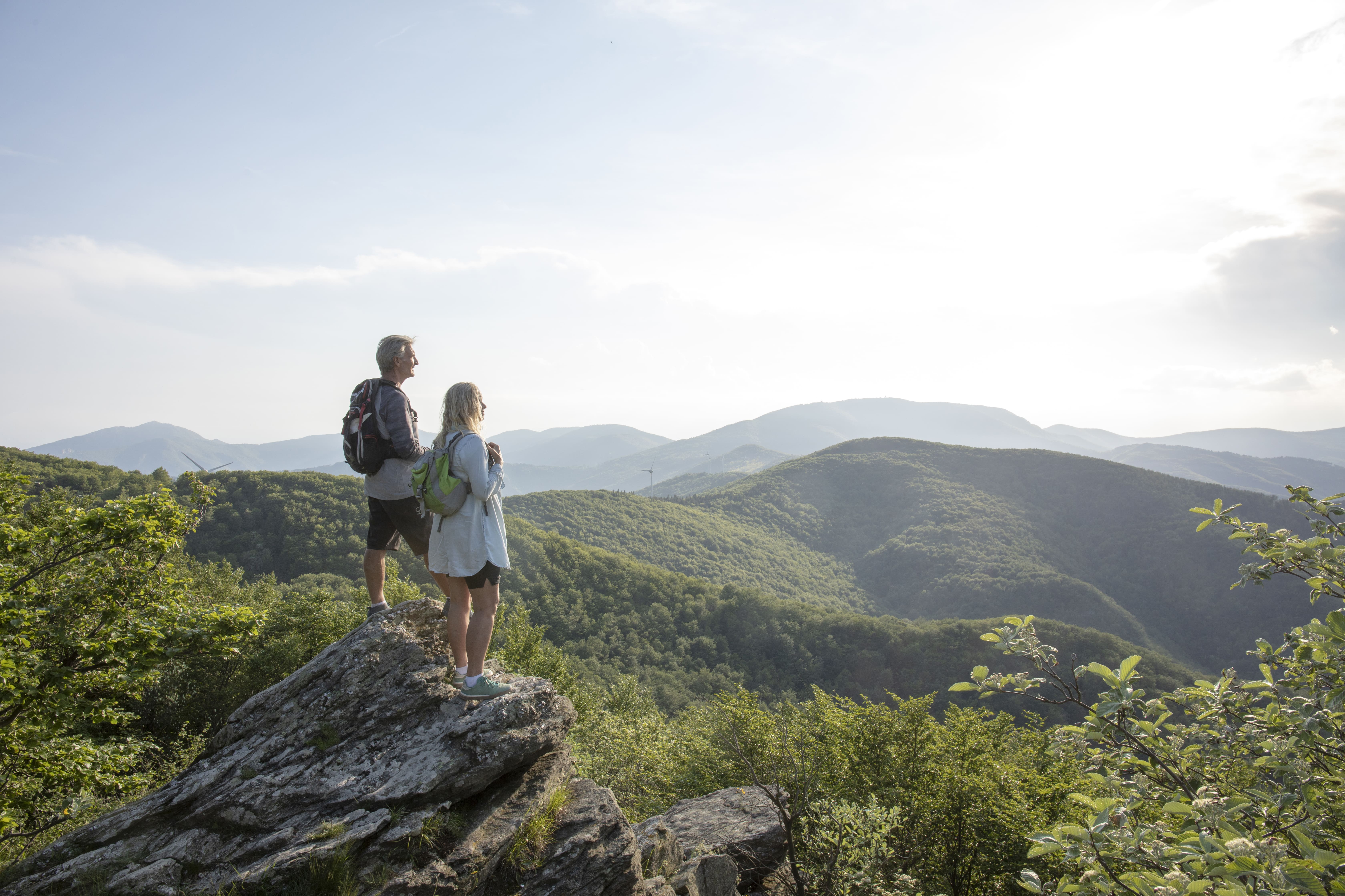 Pareja con mochilas contempla un paisaje de montañas verdes al atardecer, ideal para trekking en la naturaleza.
