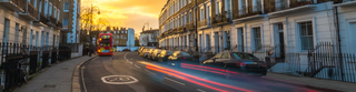 Residential London street at sunset with a car passing by and a red double decker bus pulling away from a bus stop.