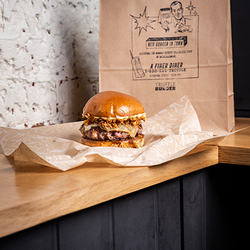 A burger placed on an open wrapper in front of a paper delivery bag.