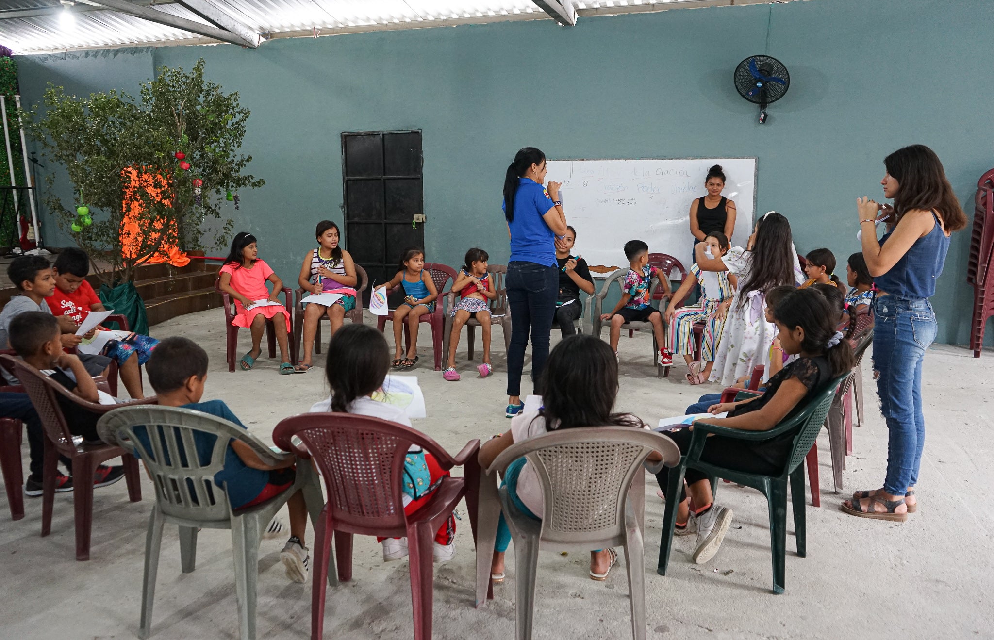 A group of children sit on chairs in a big circle with a psychologist in the centre teaching them resilience games