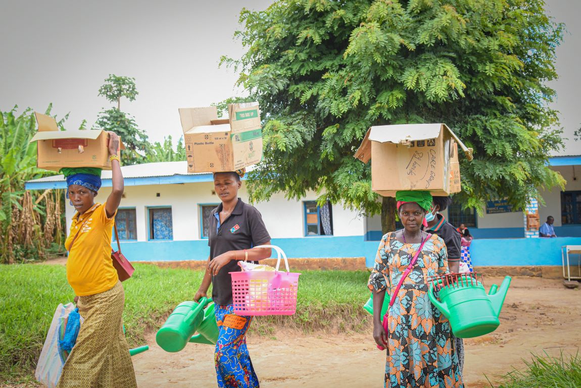 In the Democratic Republic of the Congo, three women carry cardboard boxes above their heads and in their arms carry farming and agriculture tools. 