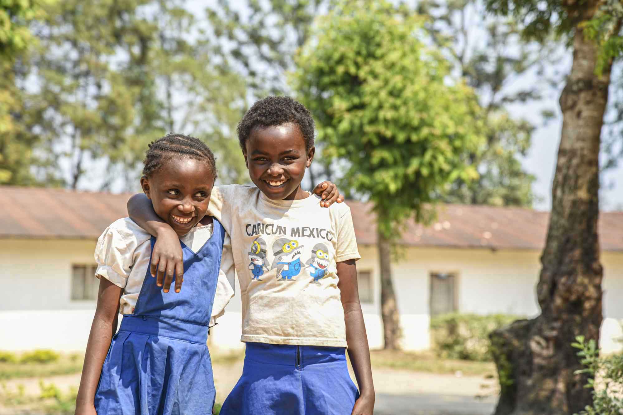 Two young children stand beside a tree, smiling and embracing each other.