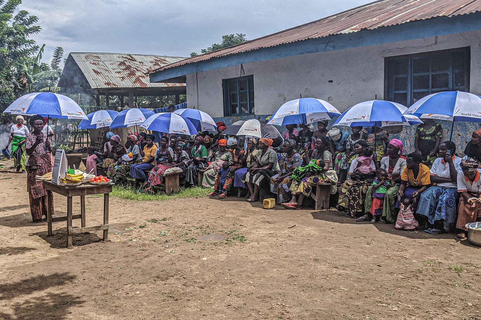 A crowd of people are seated in front of a building under blue and white umbrellas.