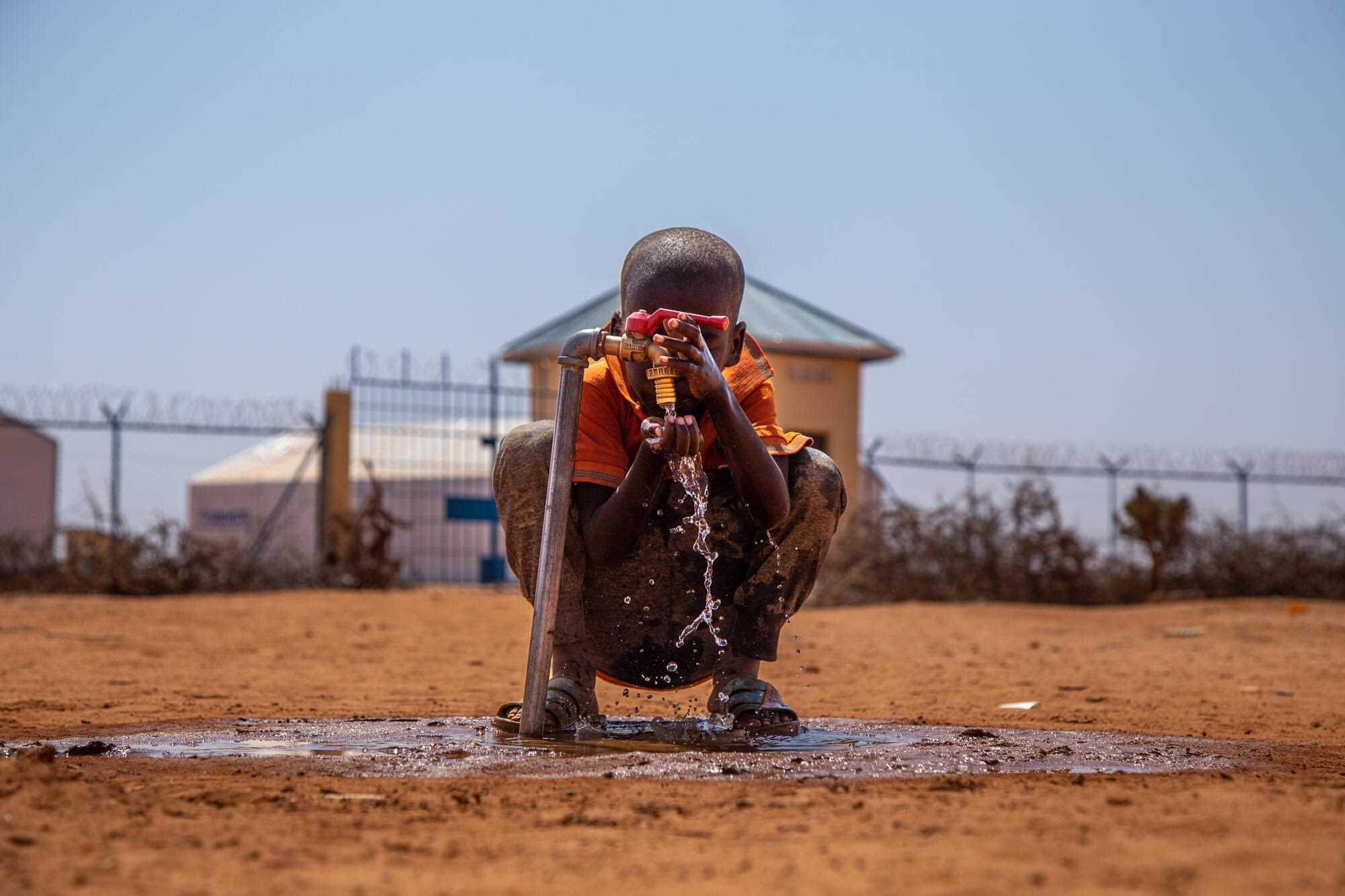In Somalia, a young boy crouches by a water tap and cups the water pouring into his hands. 