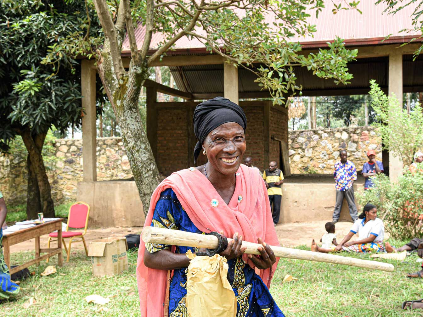 An older woman standing outside while smiling and holding a long piece of wood.
