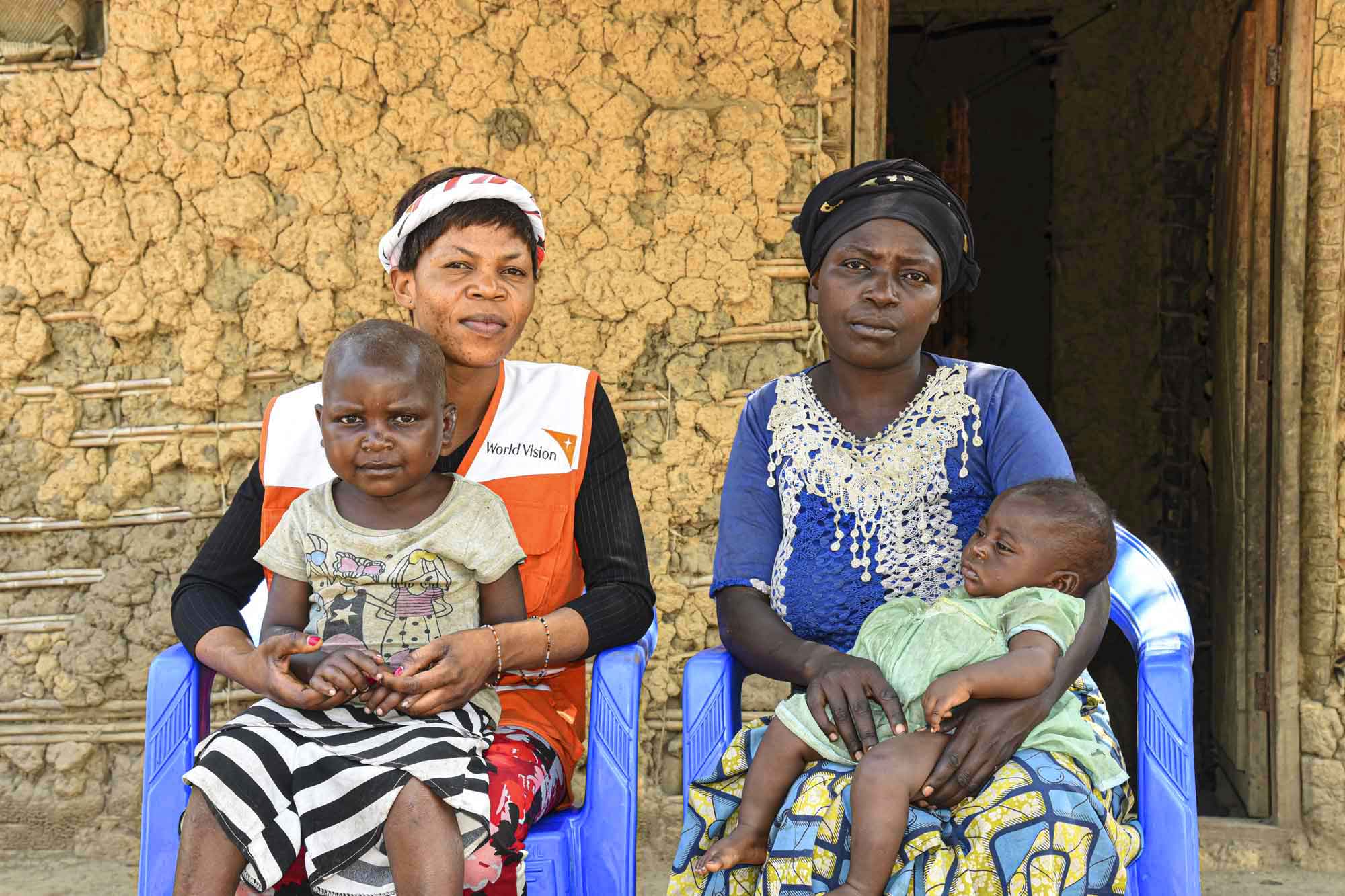 Two women are seated in front of a building on blue plastic chairs. Each are holding infant children on their laps.