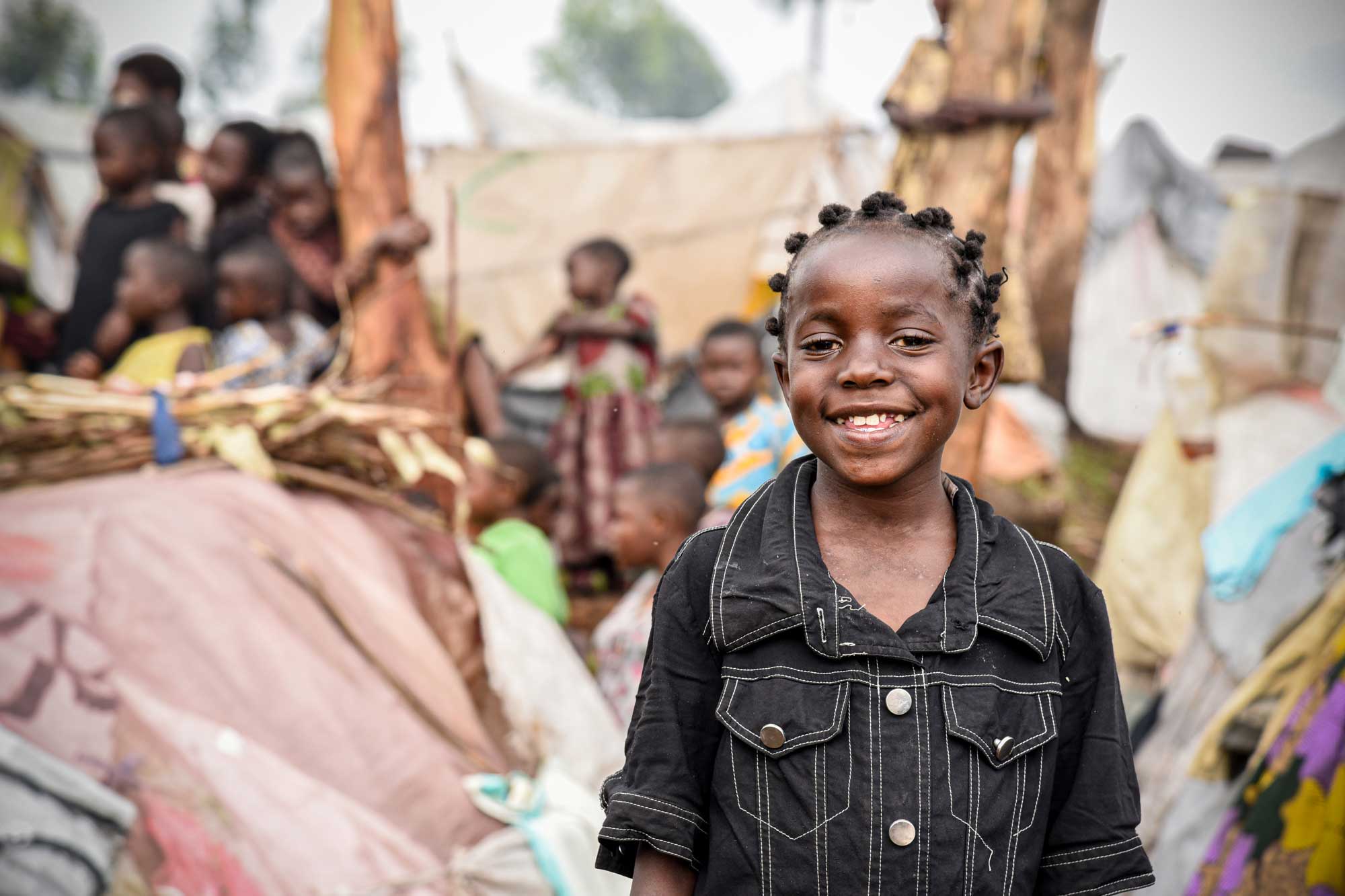 A young girl is outside wearing a denim jacket and smiling. A group of children are seen in the background.