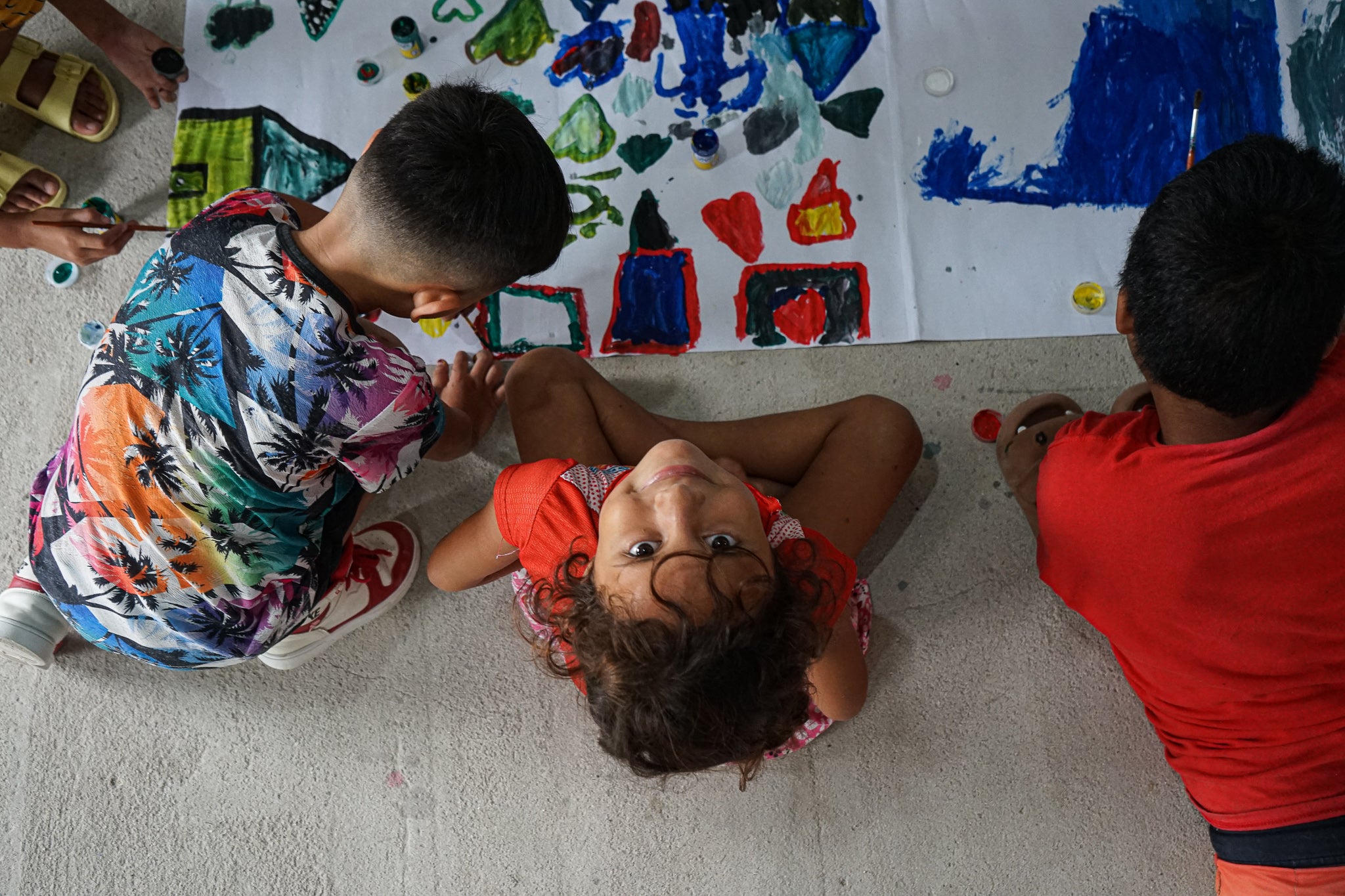 Children sit on floor doing large painting, young girl looks up to camera and smiles