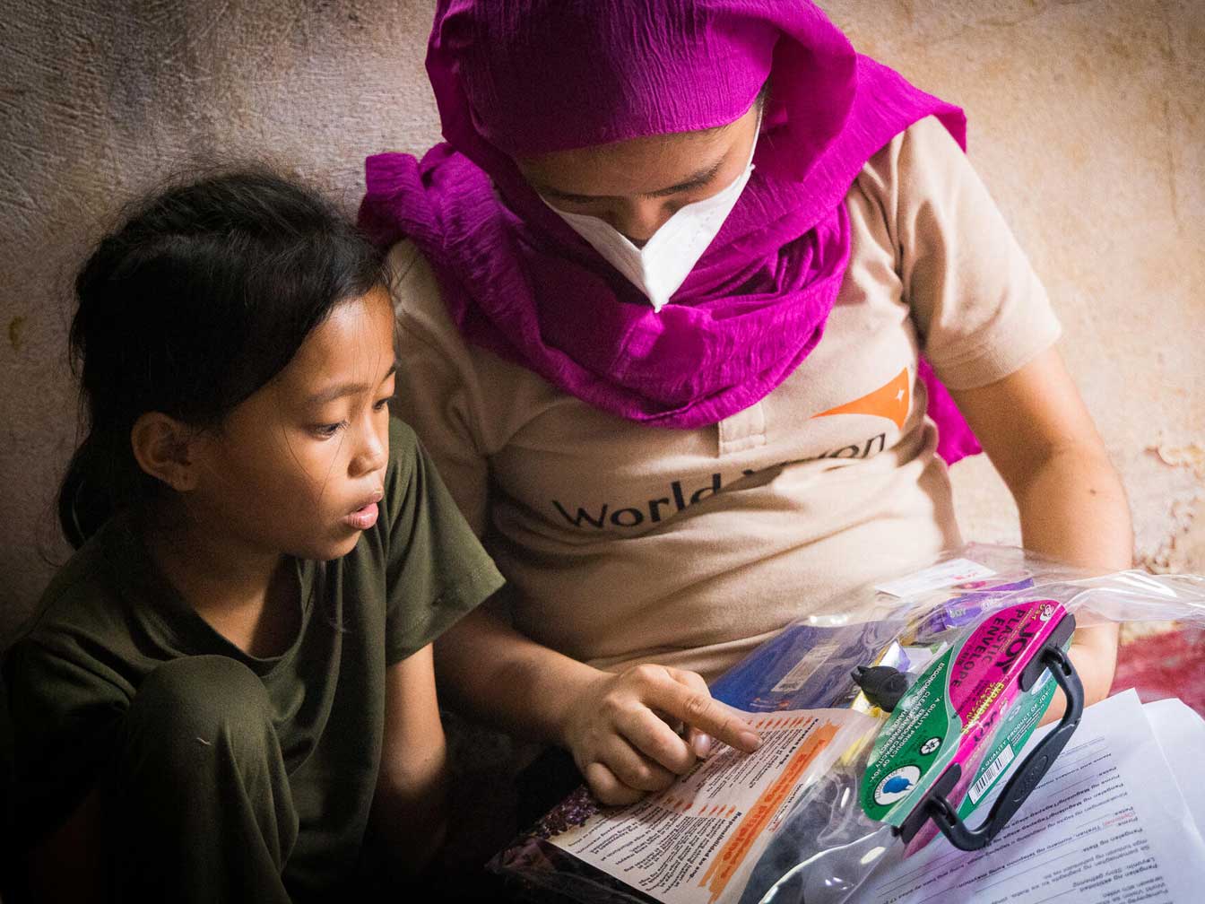 A woman in a pink scarf, reading to a child while seated on the ground.
