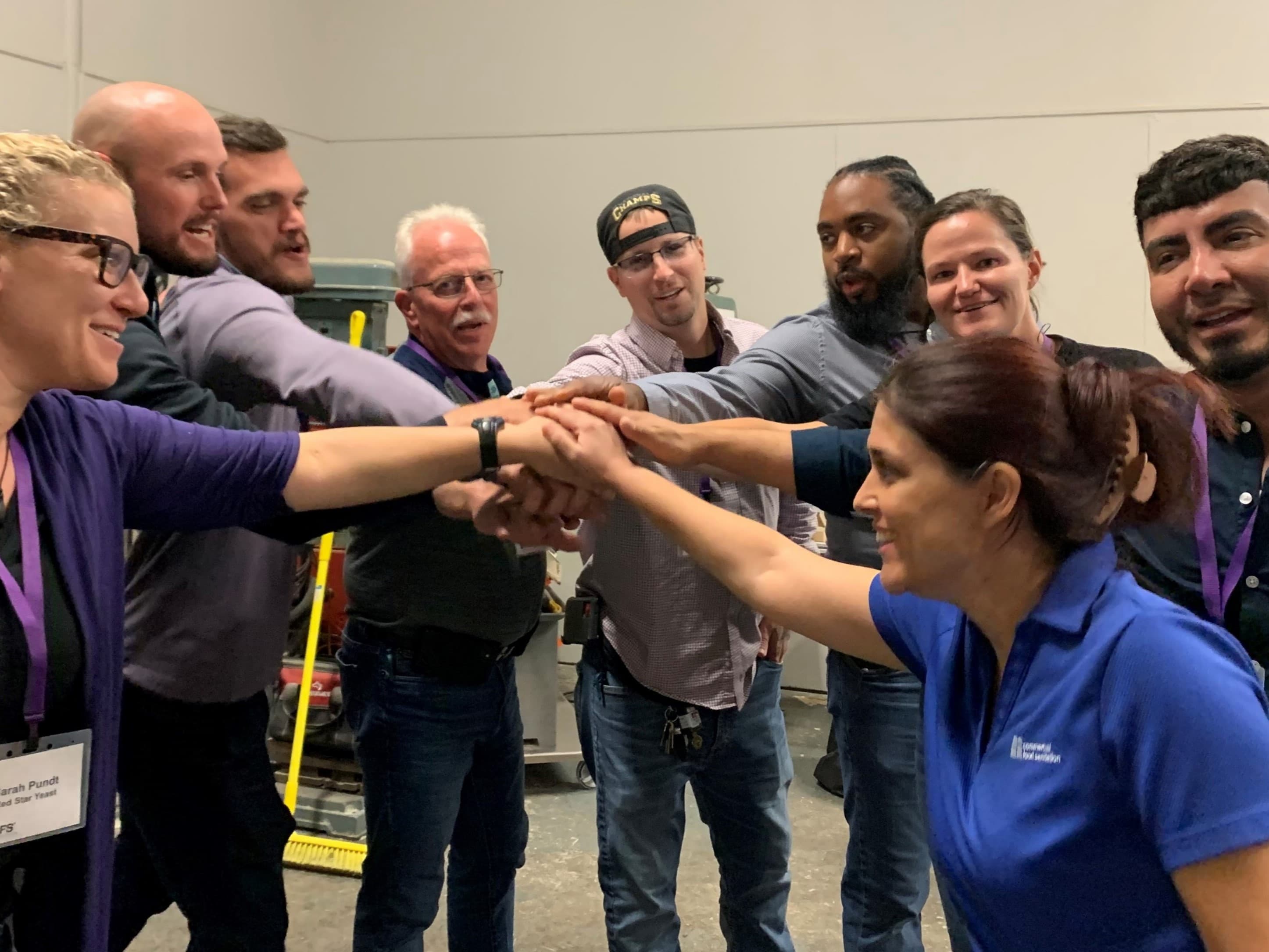A group of training participants form a close-knit circle with their hands stacked in the center. They are mid-yell, faces animated with enthusiasm and unity, embodying teamwork and camaraderie.