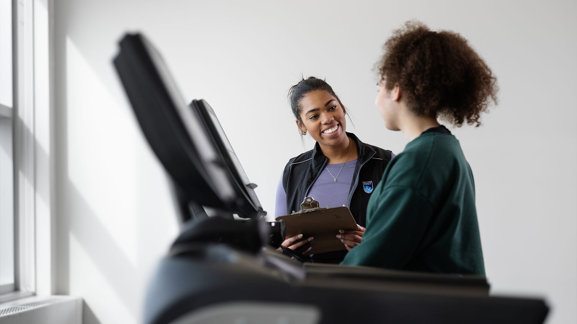 A personal fitness trainer student holds a clipboard while smiling and interacting with another student that is walking on a treadmill.
