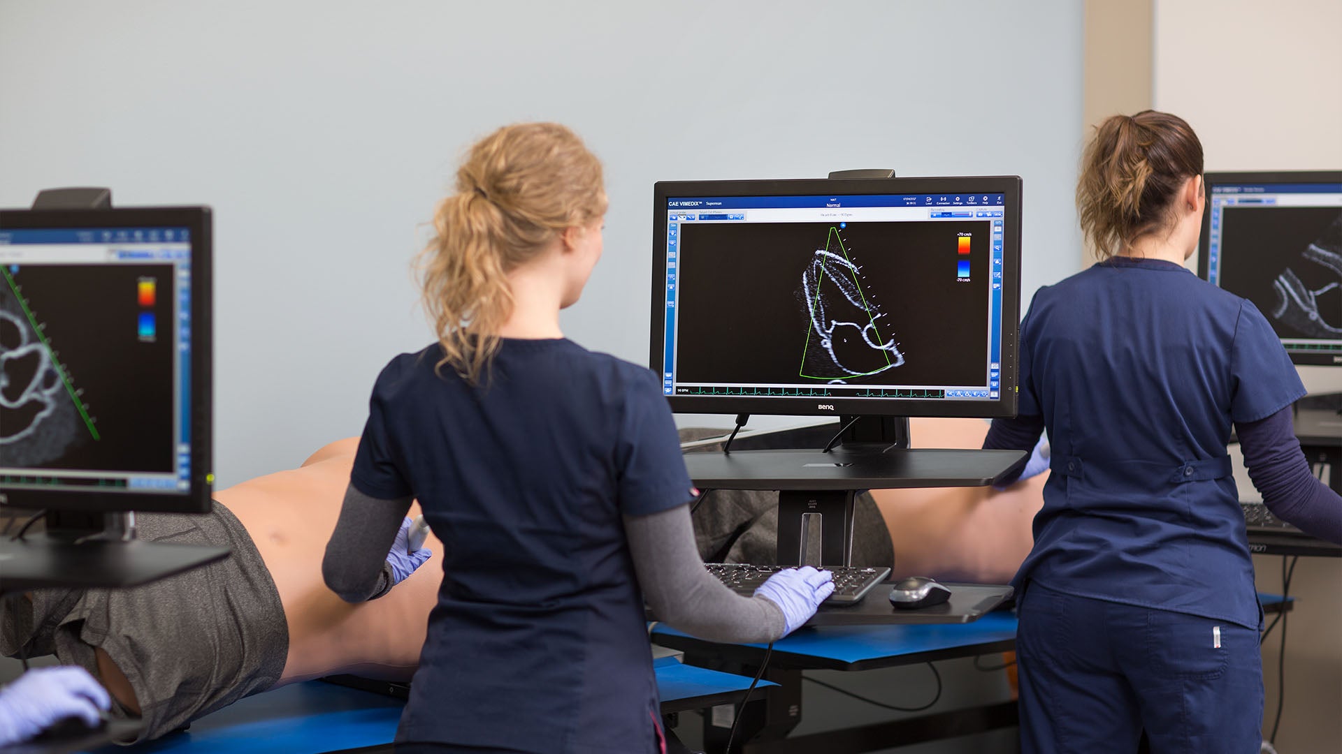 Students performing ultrasound simulations using CAE Vimedix, standing at monitors with keyboards, applying transducers to training manikins.