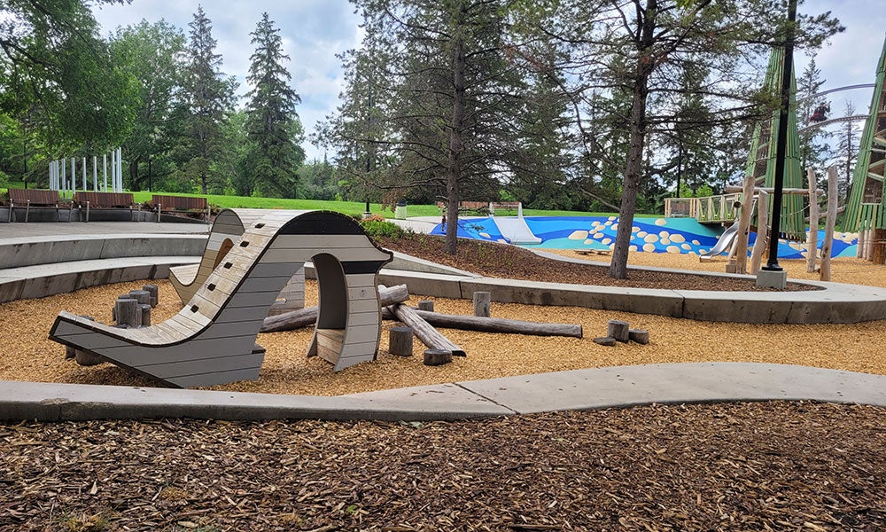 Nature-inspired playground with curved climbing structures, wood-chip surfacing, and play areas set among mature trees in a park setting.