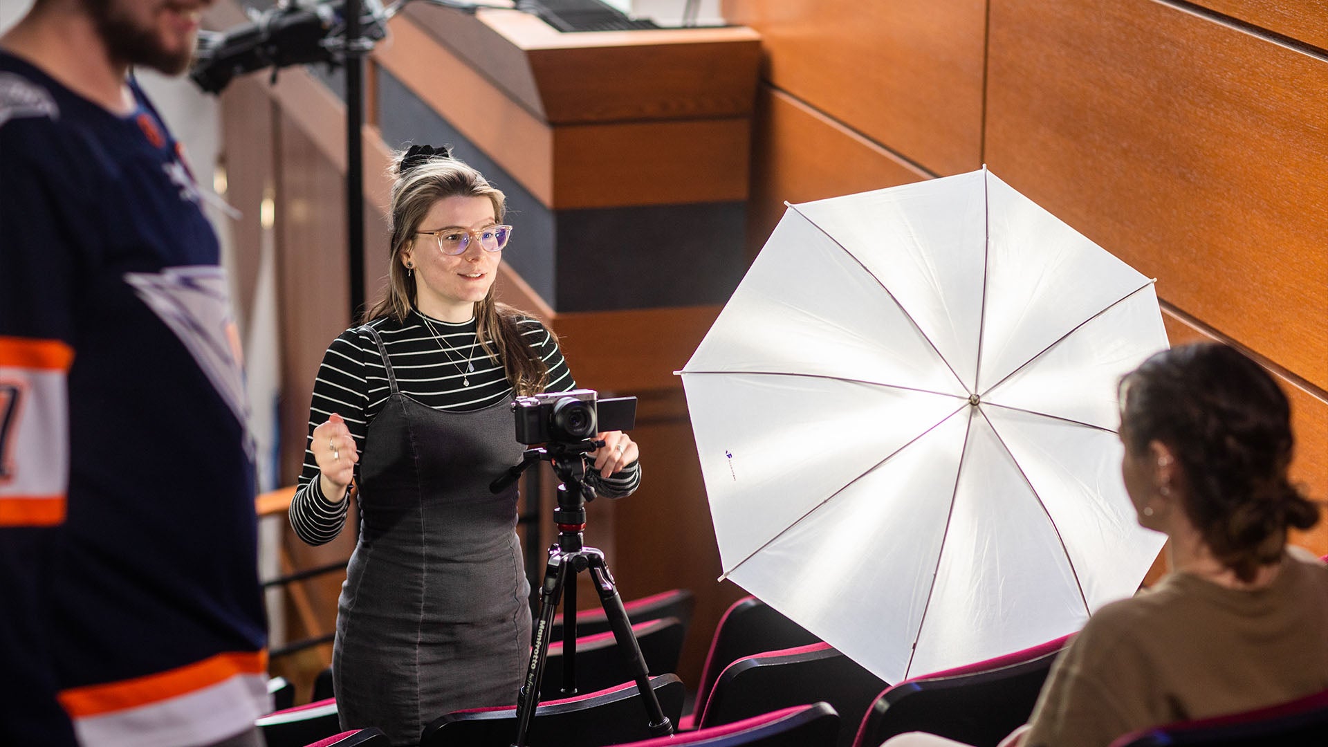 Young woman setting up camera and lighting for an interview scene in an auditorium, with a bright umbrella light and a person seated in front.