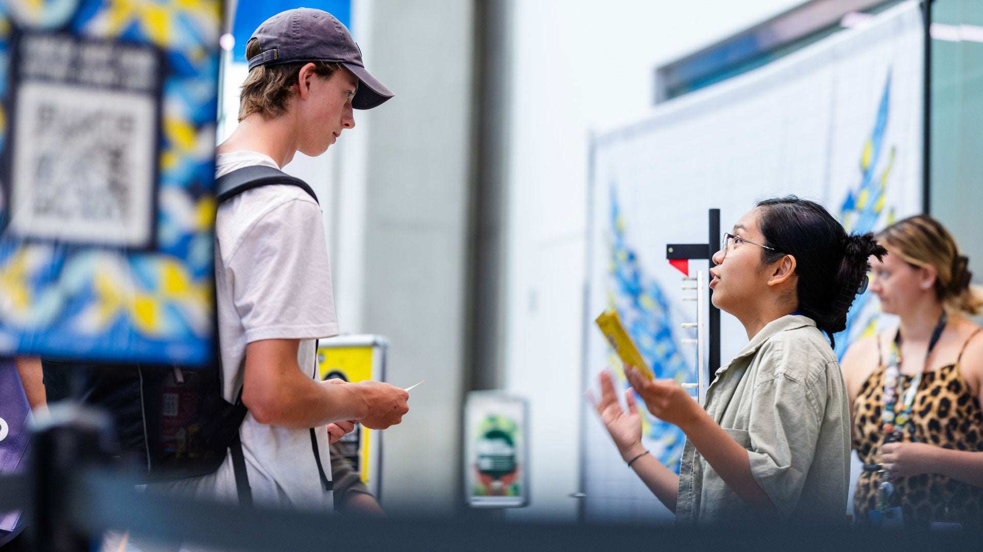 Alt text: Person speaking with another person at an information booth while holding brochures, with additional attendees in the background.
