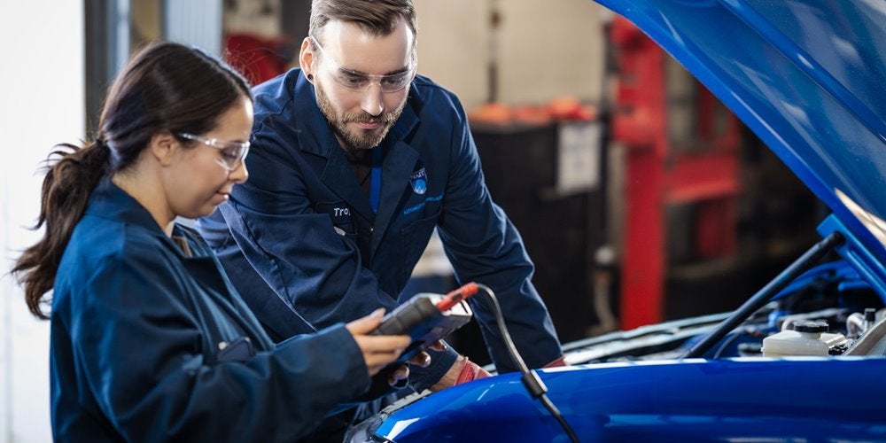 An instructor and a student wearing safety goggles and coveralls using a diagnostic tool under the hood of a car