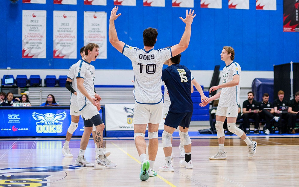 Men’s volleyball game in an indoor gymnasium, with players on the court during active play beneath championship banners and Ooks-branded athletic signage.