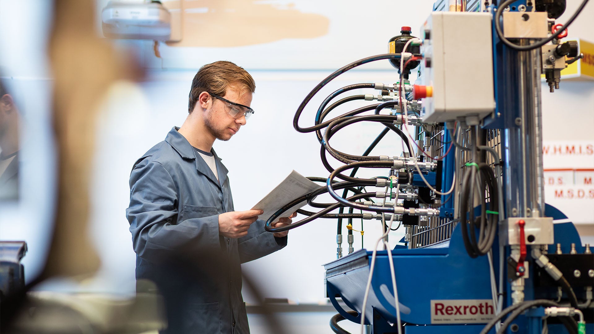 A male mechanical engineering student in a blue coat and safety glasses reads a paper while standing in front of large machinery.
