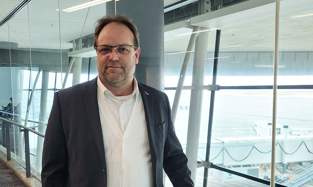 Person in a dark blazer and white shirt standing inside an airport terminal beside floor-to-ceiling windows overlooking the airfield and boarding gates.