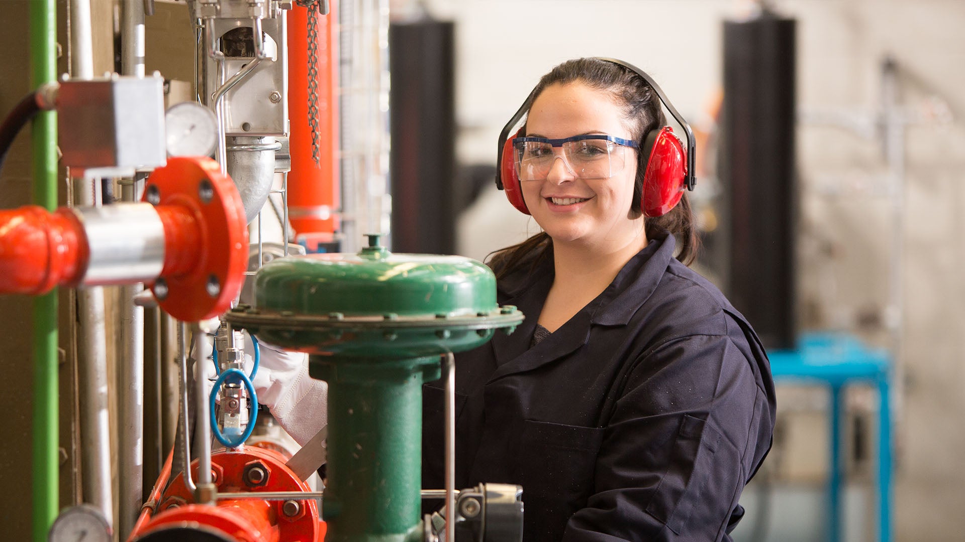 Female student wearing protective eye wear, headphones, protective gloves and navy-blue work wear, smiles while operating machinery.