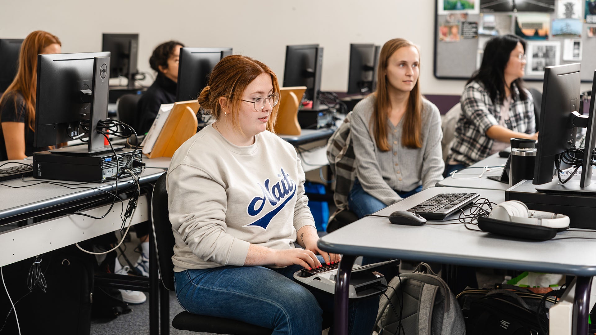 A female student sitting attentively in a classroom, using a stenotype machine, with computers and other students in the background.