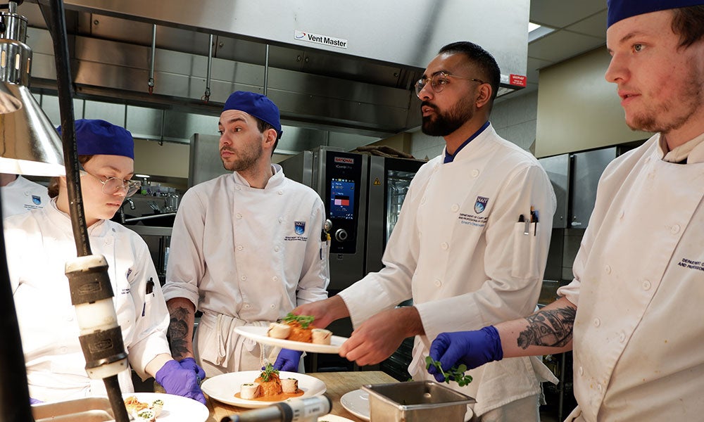 Three culinary students and an instructor in a professional kitchen plating and garnishing dishes under bright overhead lights beside stainless steel equipment.
