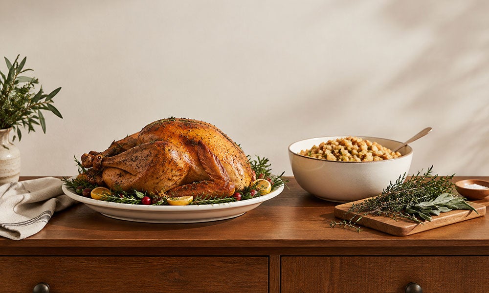 Roasted turkey on a serving platter beside a bowl of stuffing and fresh herbs on a wooden table.