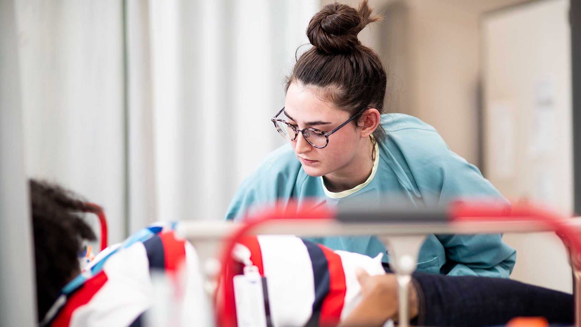 A medical lab assistant wearing a teal gown is leaning over to treat a patient who is lying down.