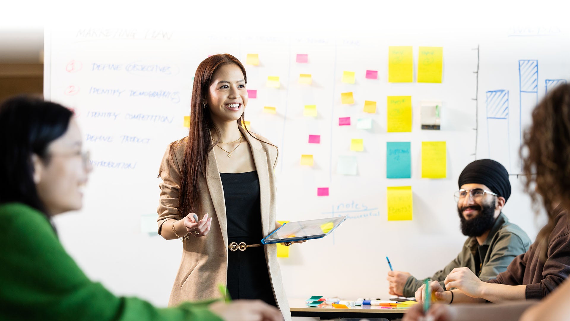 A woman holding an iPad and smiling while explaining something to a group, with a whiteboard covered with post-it notes in the background.